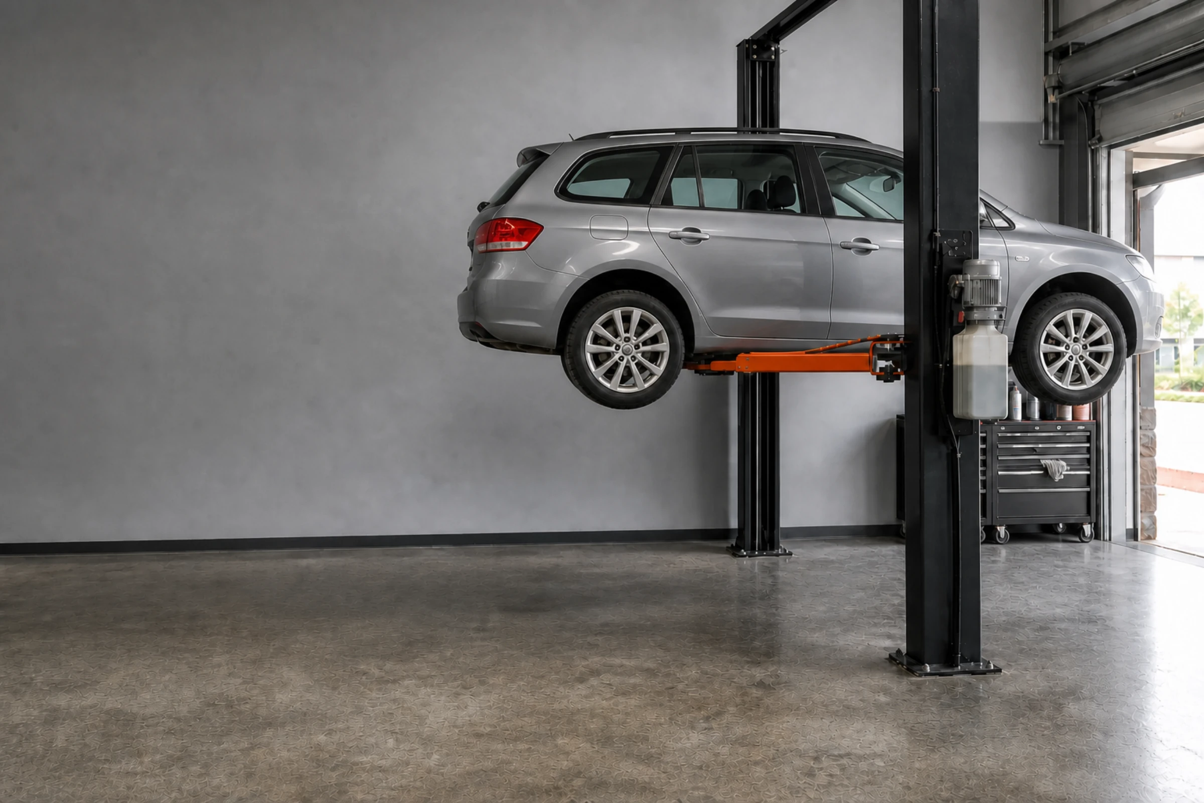 A silver car raised on a two-post lift in a two-bay workshop